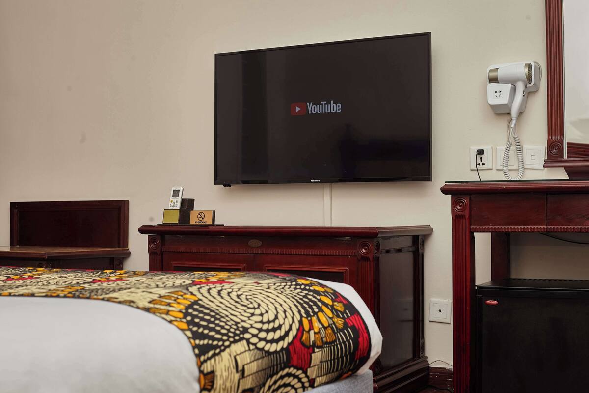 A hotel room with a bed, a TV mounted on the wall, a hairdryer on a table, and a wooden cabinet.