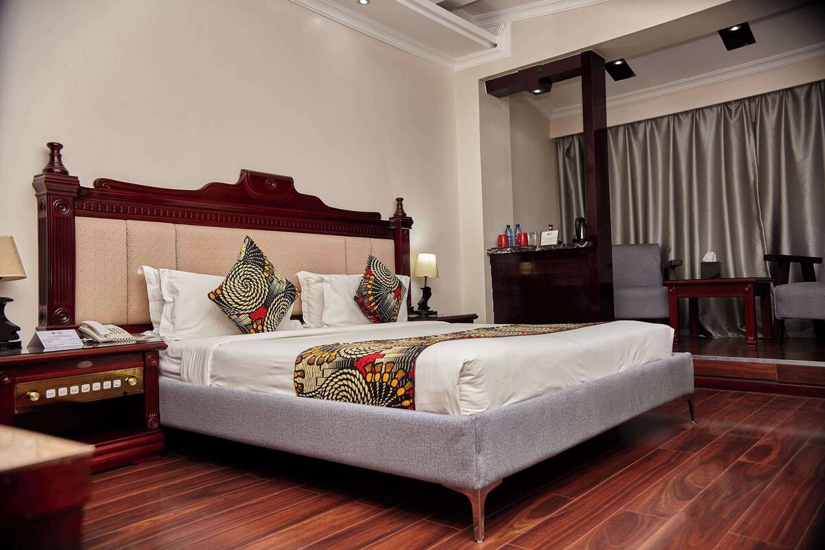 A hotel room with a large bed, a nightstand, a desk, a chair, and a view of the outside through a window.