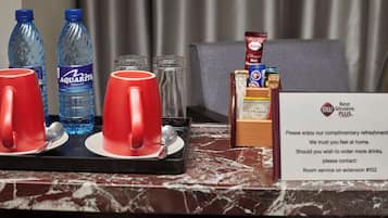 Un espace d’accueil d’hôtel avec un comptoir en marbre, deux bouteilles d’eau Aquarite, deux tasses rouges, un verre, une cuillère, un sachet de chocolat chaud Slojo et un panneau indiquant que des boissons sont offertes gratuitement.