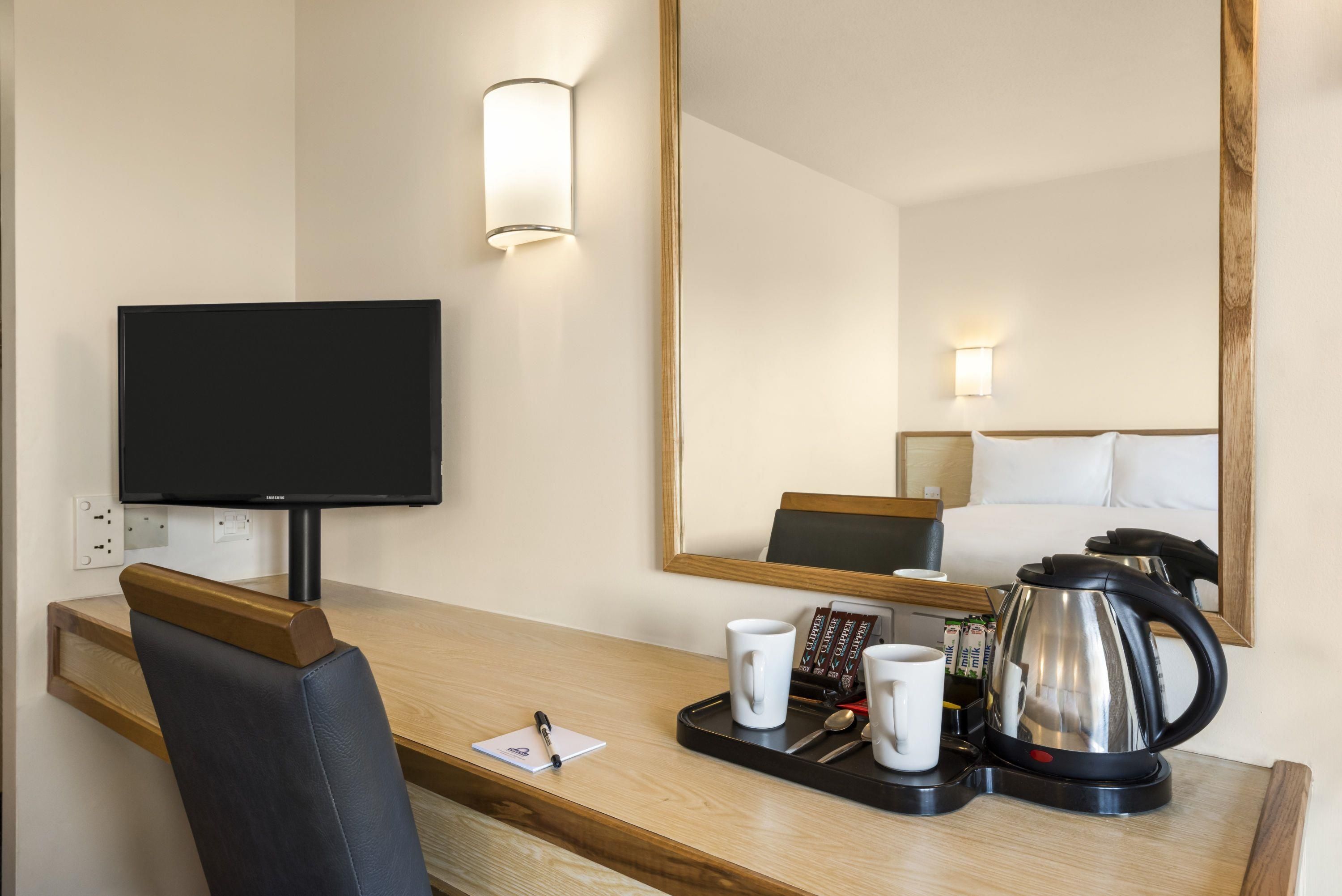 A hotel room with a flat-screen TV, a wooden desk, a coffee maker, and a tray with cups and a pen.