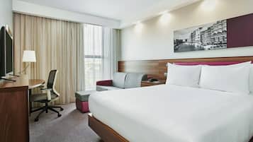 A hotel room with a large bed, a desk with a chair, a small seating area, and a large window with curtains.