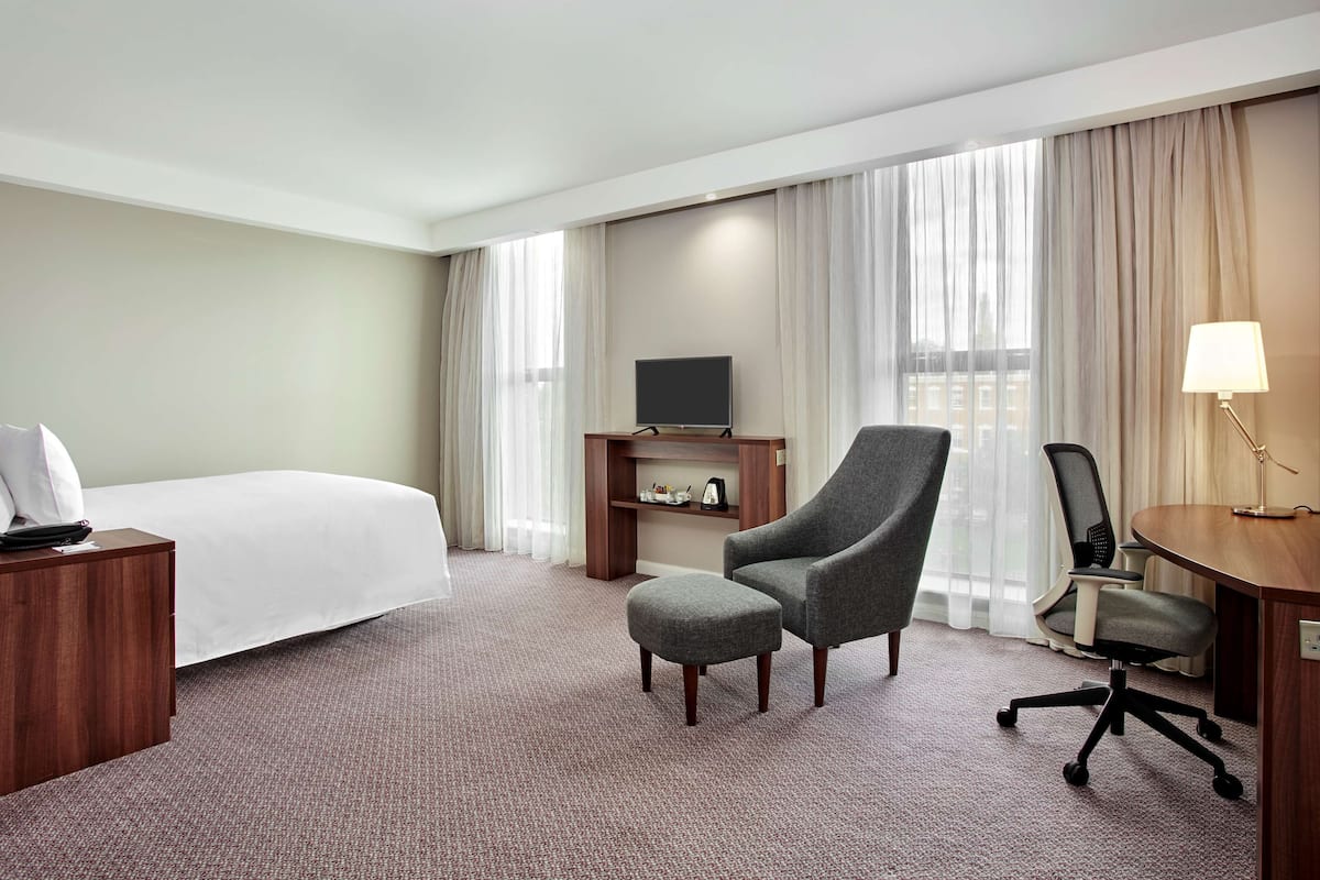 A hotel room with a bed, a grey armchair, a desk with a lamp, a TV on a wooden stand, and a view of the city through large windows.