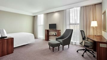 A hotel room with a bed, a grey armchair, a desk with a lamp, a TV on a wooden stand, and a view of the city through large windows.
