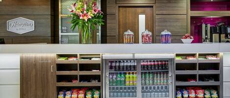 Zona de recepción de un hotel con un mostrador de madera, un arreglo floral grande y una vitrina refrigerada para bebidas.