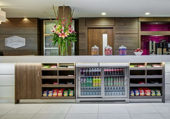 Un espace d’accueil d’hôtel avec un comptoir de réception en bois, un grand arrangement floral et un présentoir réfrigéré pour les boissons.