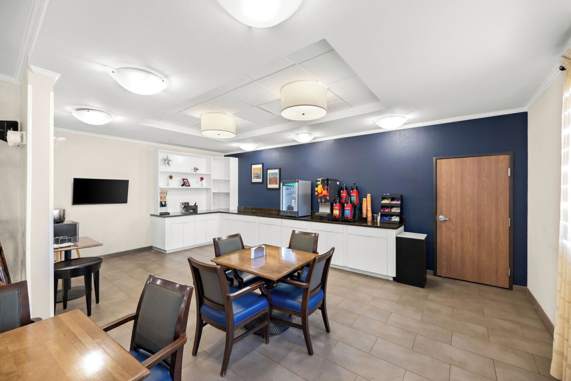A modern kitchen with white cabinets, a dining area with wooden tables and chairs, a refrigerator, and a coffee machine.