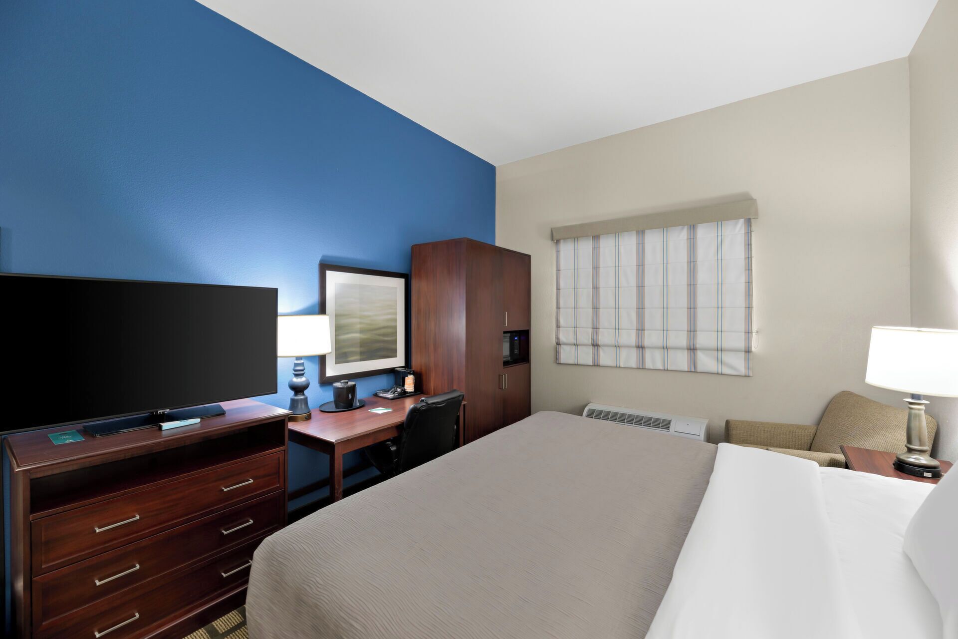 A hotel room with a large bed, a desk with a computer, a TV, and a window with blinds.
