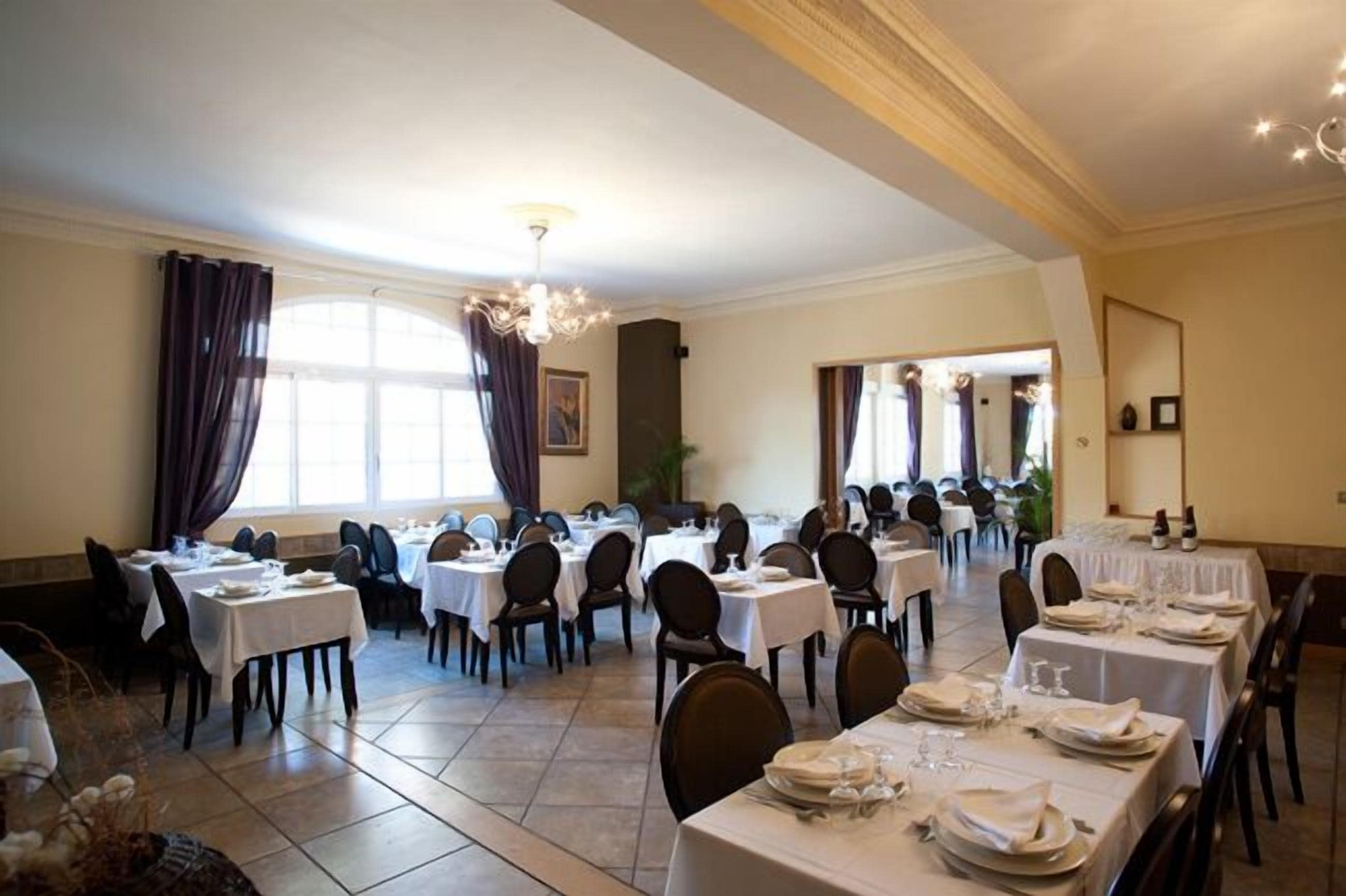 A large dining hall with multiple tables set for dining, featuring a chandelier and large windows.