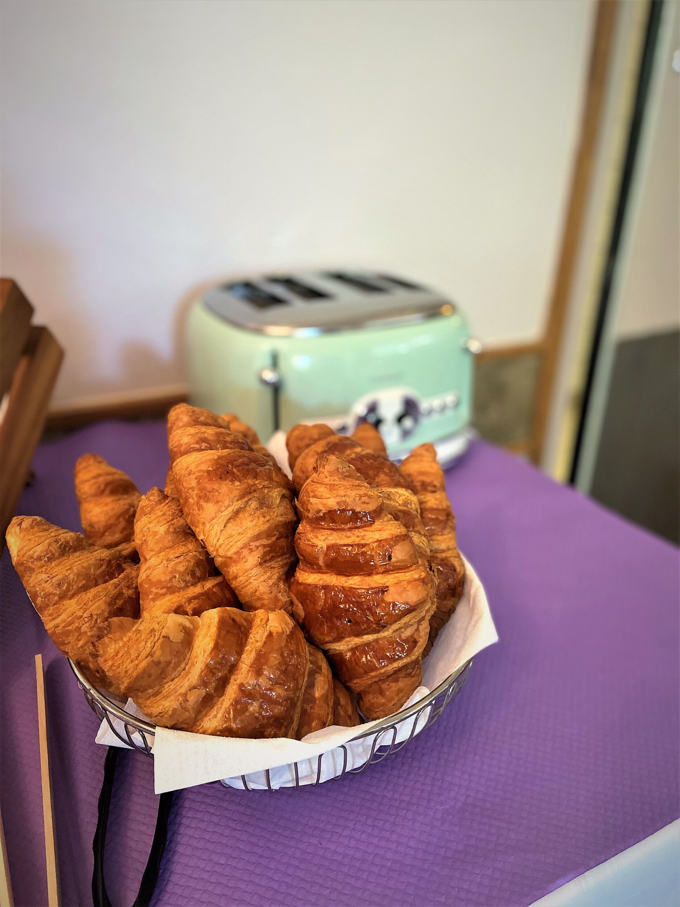 A basket of croissants on a purple tablecloth with a vintage toaster in the background.