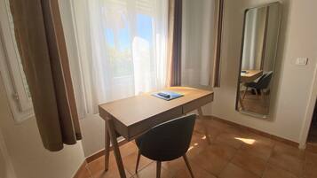 Um quarto pequeno com escrivaninha de madeira, uma cadeira, um espelho e uma janela com cortinas.