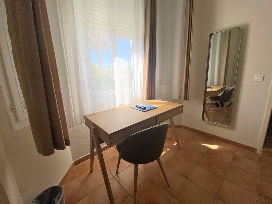 Um quarto pequeno com escrivaninha de madeira, uma cadeira, um espelho e uma janela com cortinas.