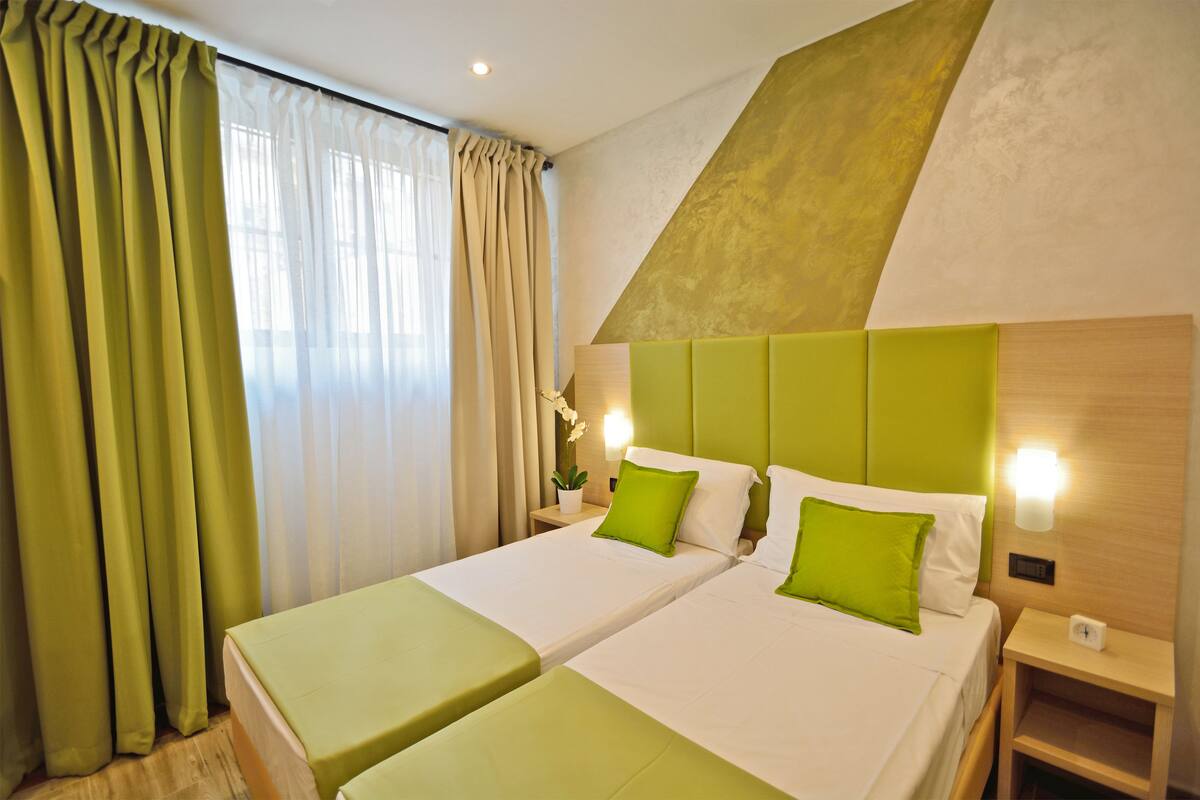Hotelkamer met twee bedden, een groene accentmuur, een nachtkastje met een lamp en een raam met gordijnen.