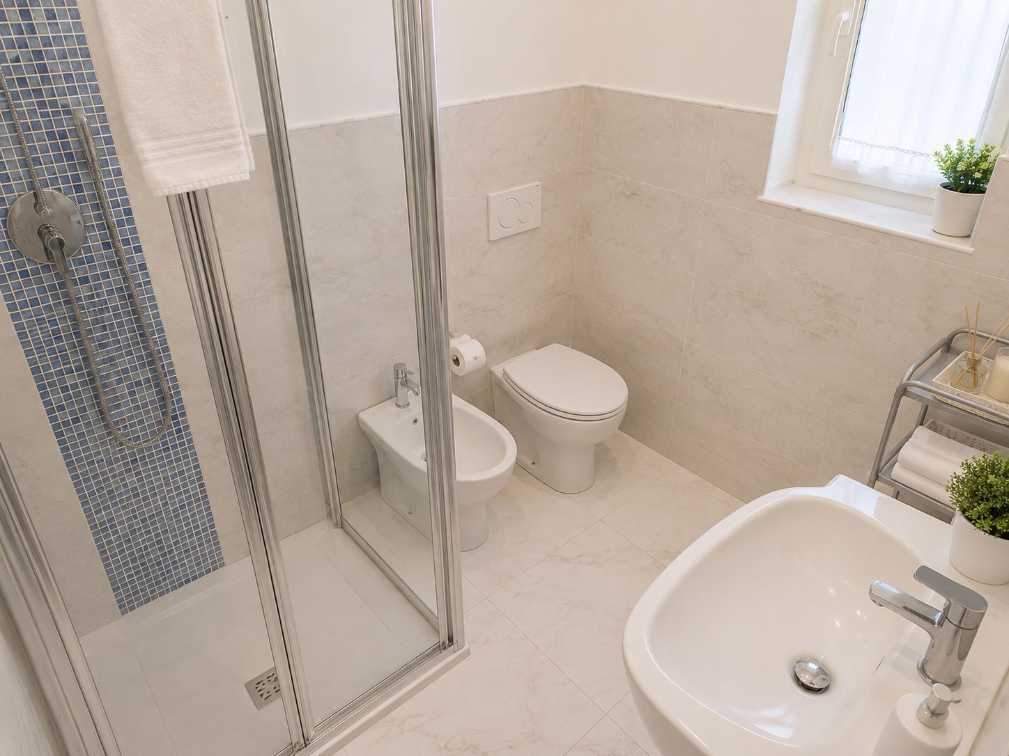 A bathroom with a glass shower enclosure, a toilet, a bidet, a sink, and a window with a plant.