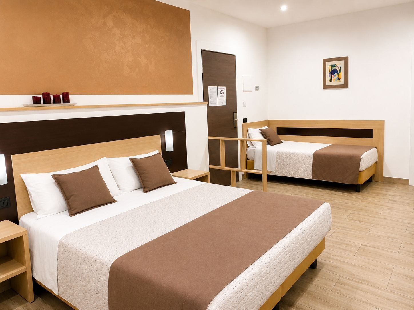 Une chambre d’hôtel avec un grand lit, un lit plus petit, une table de chevet, une tête de lit en bois, une étagère fixée au mur et une porte.