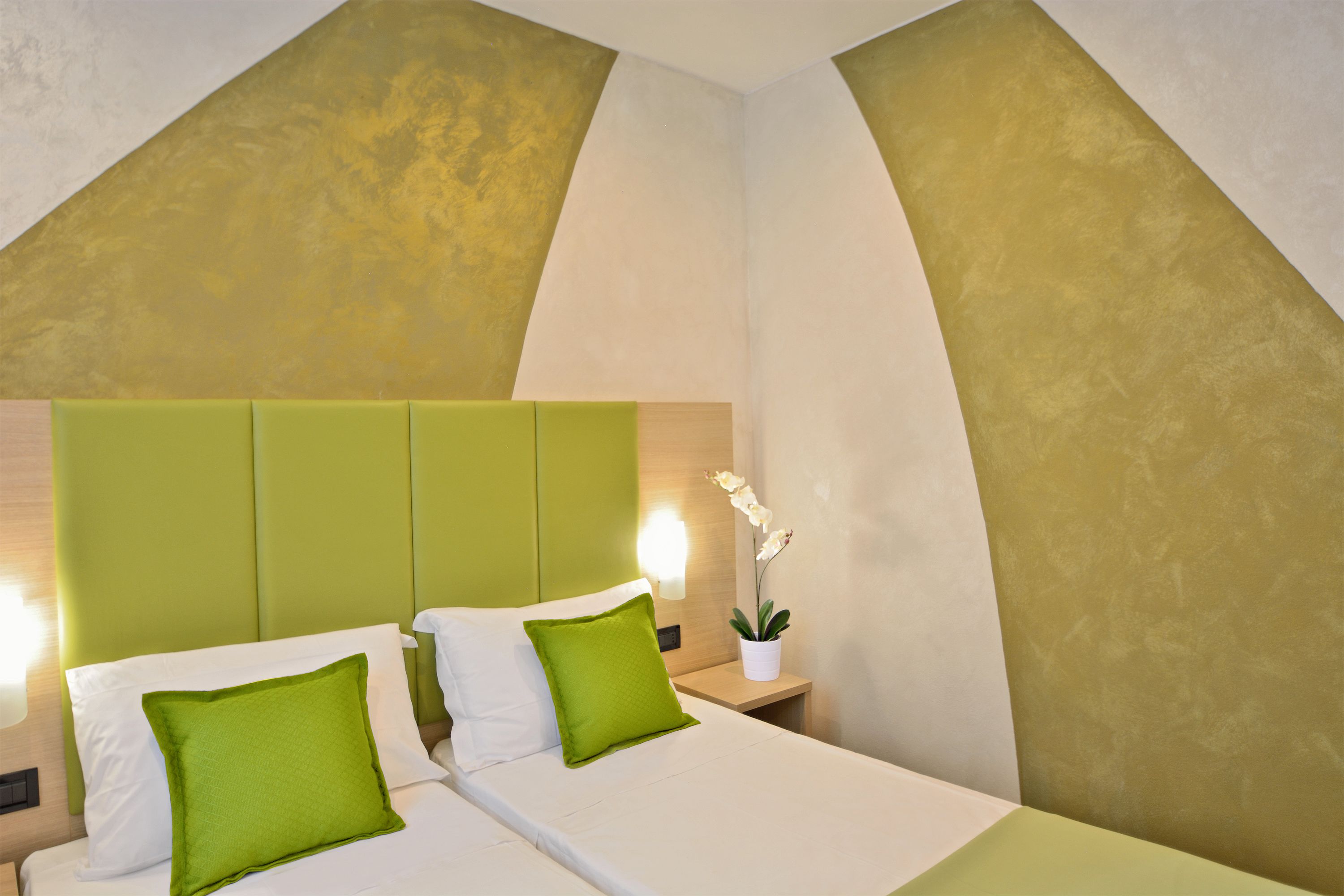 A modern hotel room with a green accent wall, a large bed with white and green pillows, a bedside table with a lamp, and a potted plant.
