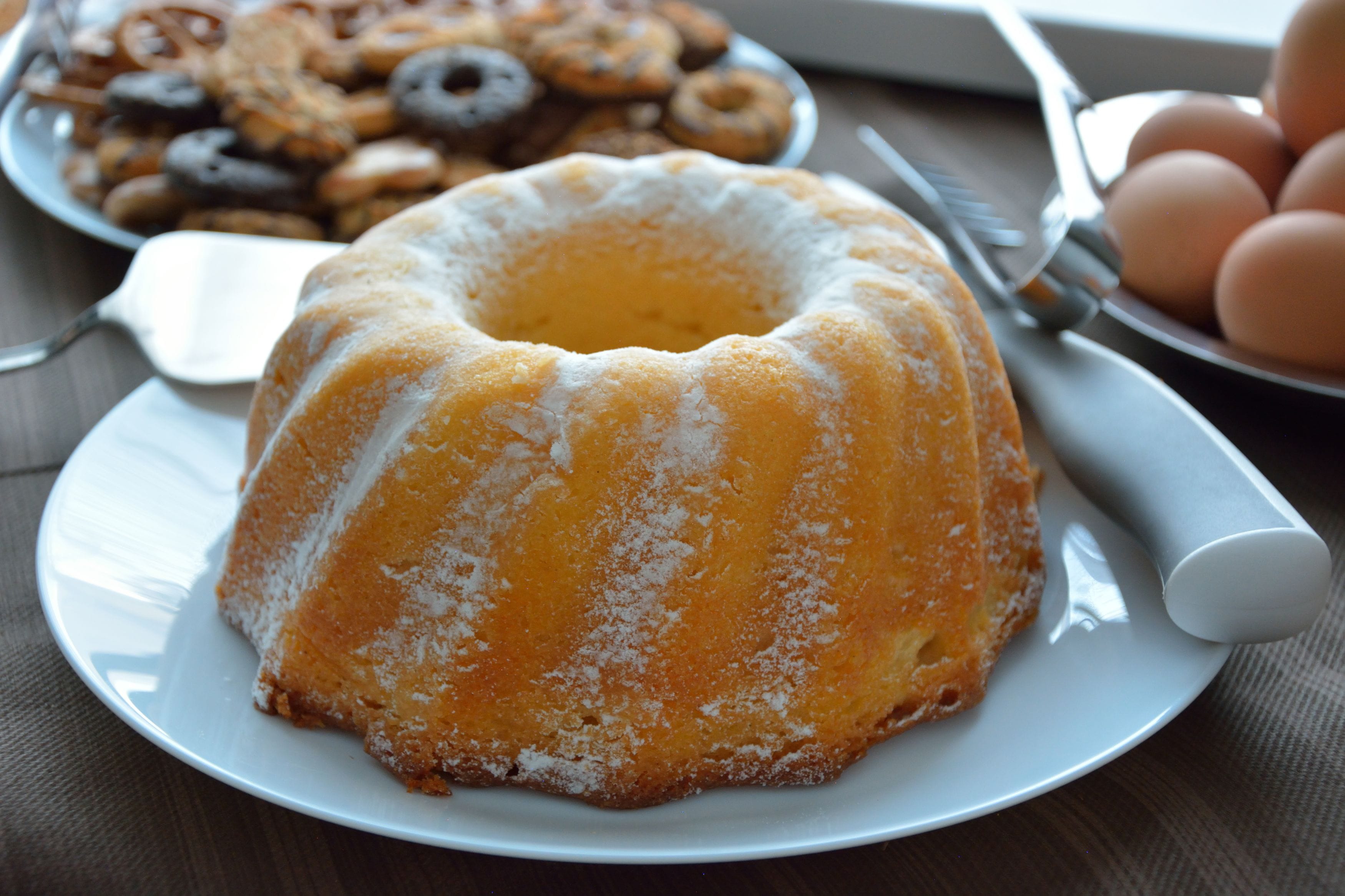A bundt cake on a plate with a spatula and a plate of donuts in the background.
