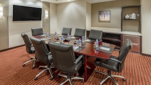 A conference room with a large table, six chairs, a flat-screen TV, and a shelf with decorative items.