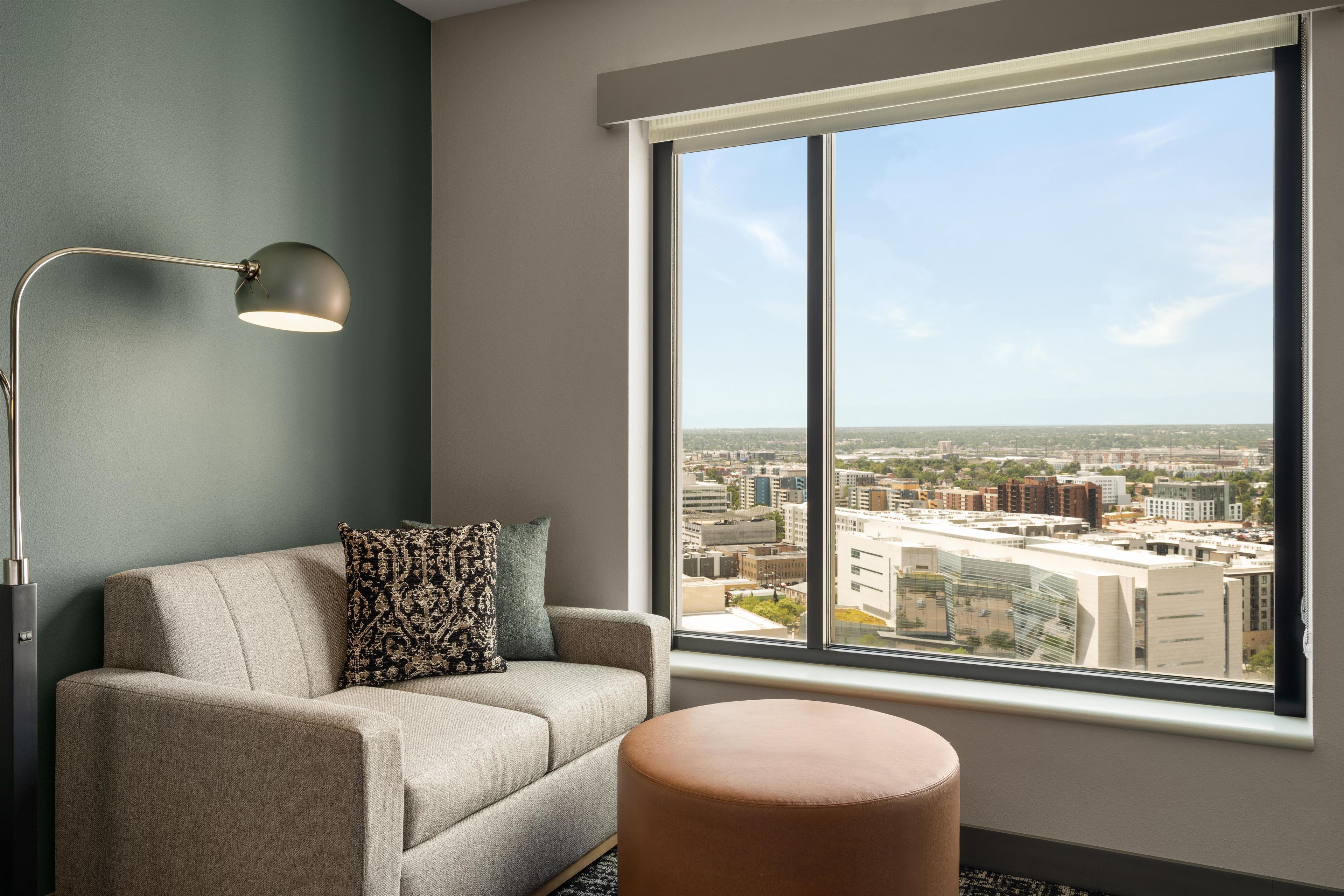 Un salon moderne avec un canapé, un pouf rond, une lampe sur pied et une grande fenêtre donnant sur la ville.