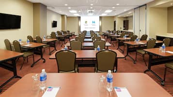 Un salón de conferencias con varias mesas y sillas, botellas de agua y una pantalla para presentaciones.