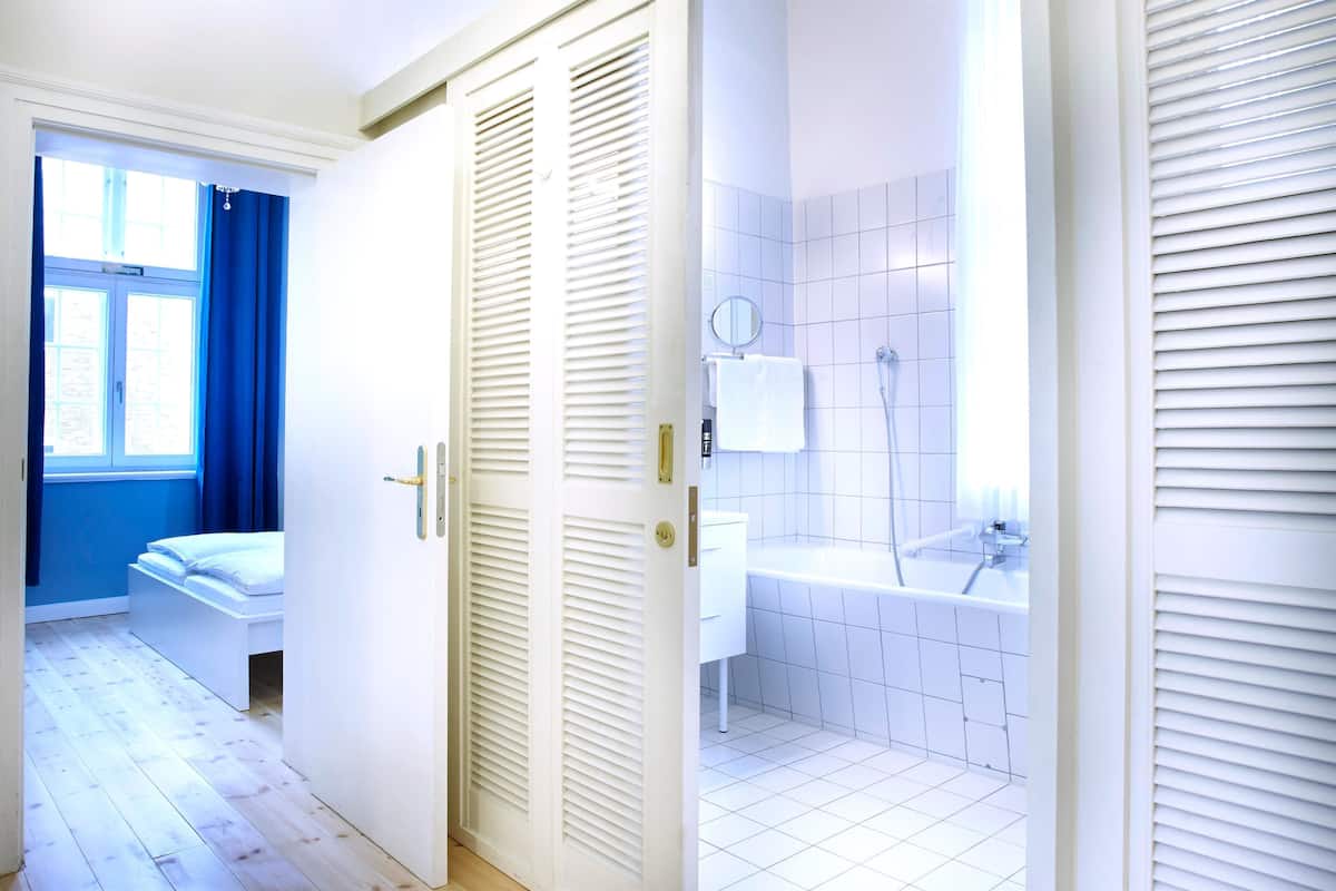 A hotel room with a white bed, a white bathroom with a bathtub, and a window with blue curtains.