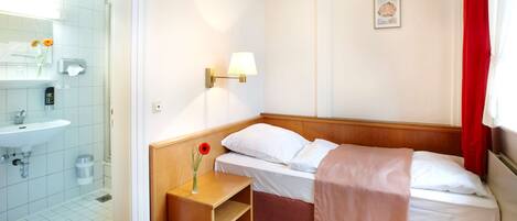 Habitación de hotel con cama de madera, ropa de cama blanca, lámpara de noche, mesita de noche de madera con un florero y baño con lavamanos y espejo.
