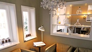 Een gezellige woonkamer met twee fauteuils, een bijzettafeltje met een lamp, een raam met een plank vol kaarsen, en een spiegel die een eethoek weerkaatst.