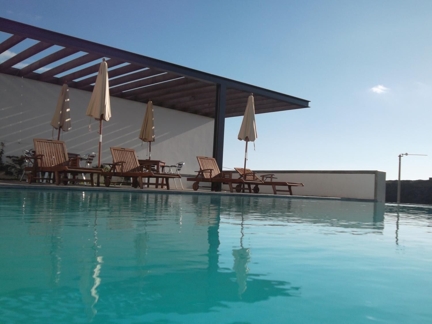 A swimming pool with lounge chairs and umbrellas, a covered seating area, and a clear sky.