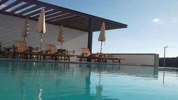A swimming pool with lounge chairs and umbrellas, a covered seating area, and a clear sky.