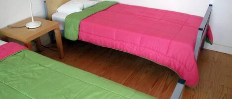 Une chambre avec deux lits simples, l’un recouvert d’une couvre-lit verte et l’autre d’une couvre-lit rose. Une table de chevet en bois avec une lampe blanche se trouve à côté du lit vert.