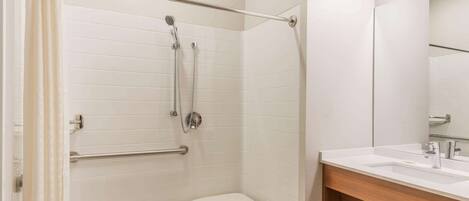 A bathroom with a shower, a white tiled wall, a wooden vanity with a mirror, and a white bench.