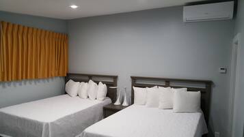 Habitación de hotel con dos camas individuales, una mesita de noche, aire acondicionado instalado en la pared y una ventana con cortinas amarillas.