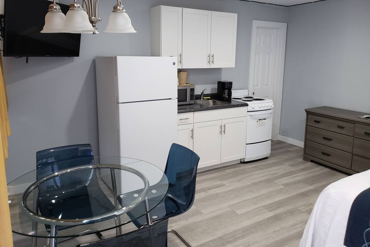 A compact kitchen with white cabinets, a stove, and a microwave. There is a glass table with chairs and a wooden cabinet to the right.