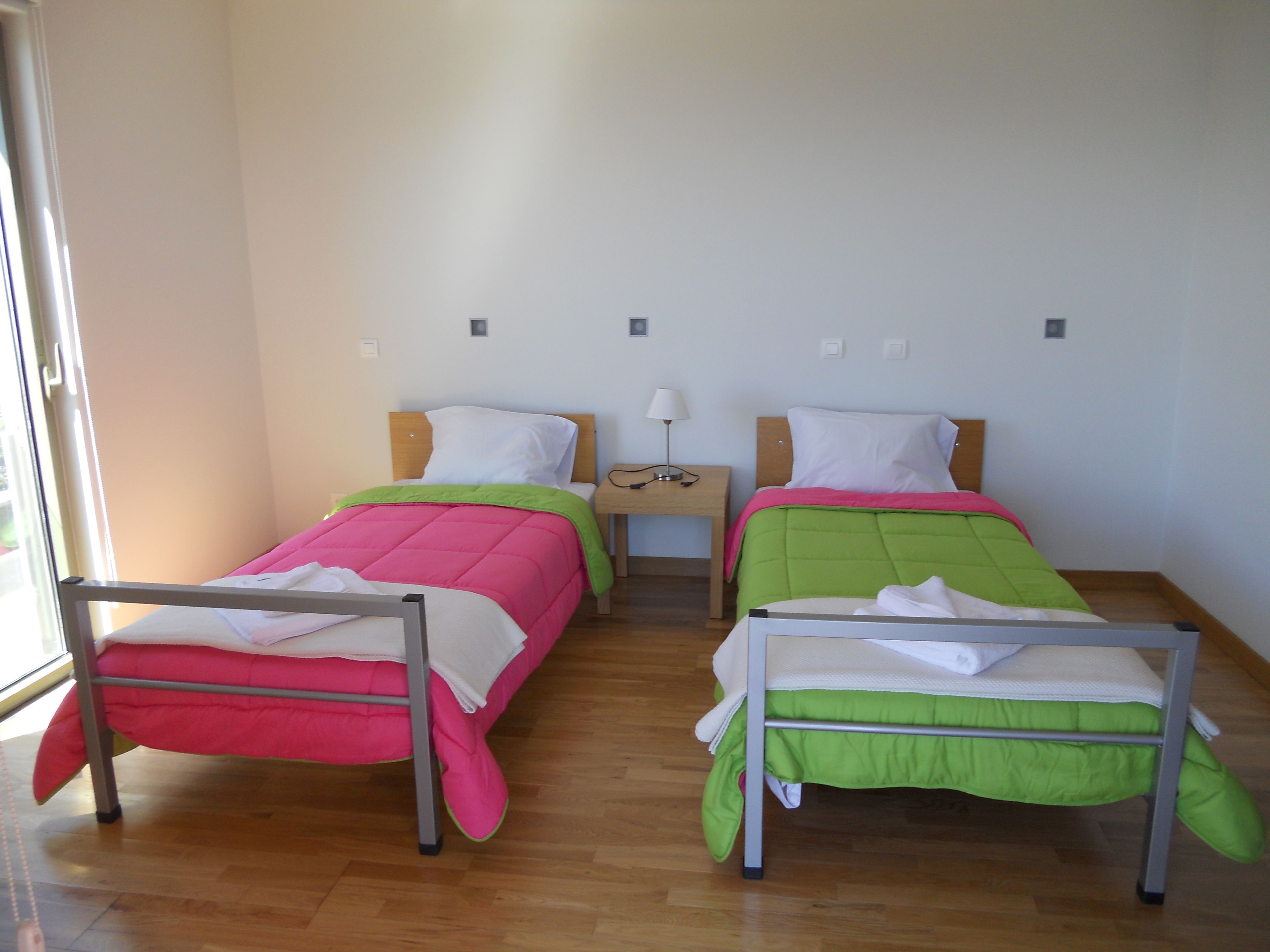 Een kamer met twee eenpersoonsbedden, elk met een uniek gekleurd sprei, een nachtkastje met een lamp, en een raam dat natuurlijk licht binnenlaat.