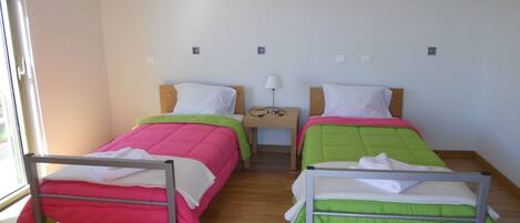 Een kamer met twee eenpersoonsbedden, elk met een uniek gekleurd sprei, een nachtkastje met een lamp, en een raam dat natuurlijk licht binnenlaat.