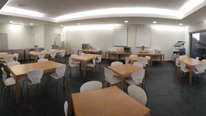 A modern conference room with multiple tables and chairs, equipped with a whiteboard, a coffee machine, and a window offering a view of the sea. - Azores Youth Hostels - São Jorge (Calheta)