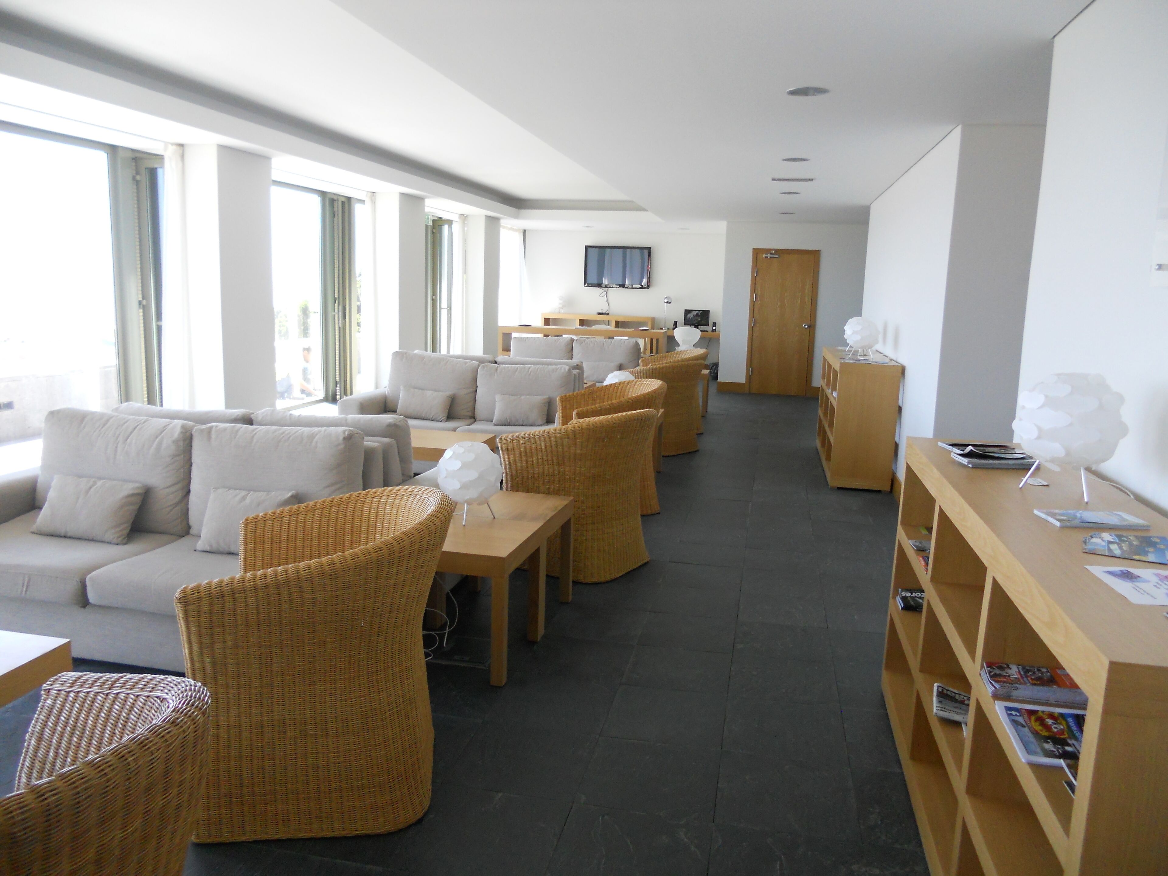 Eine moderne Hotellobby mit einer langen Rezeption, mehreren Sitzbereichen mit Sofas und Korbstühlen sowie einem Holzregal mit Zeitschriften.