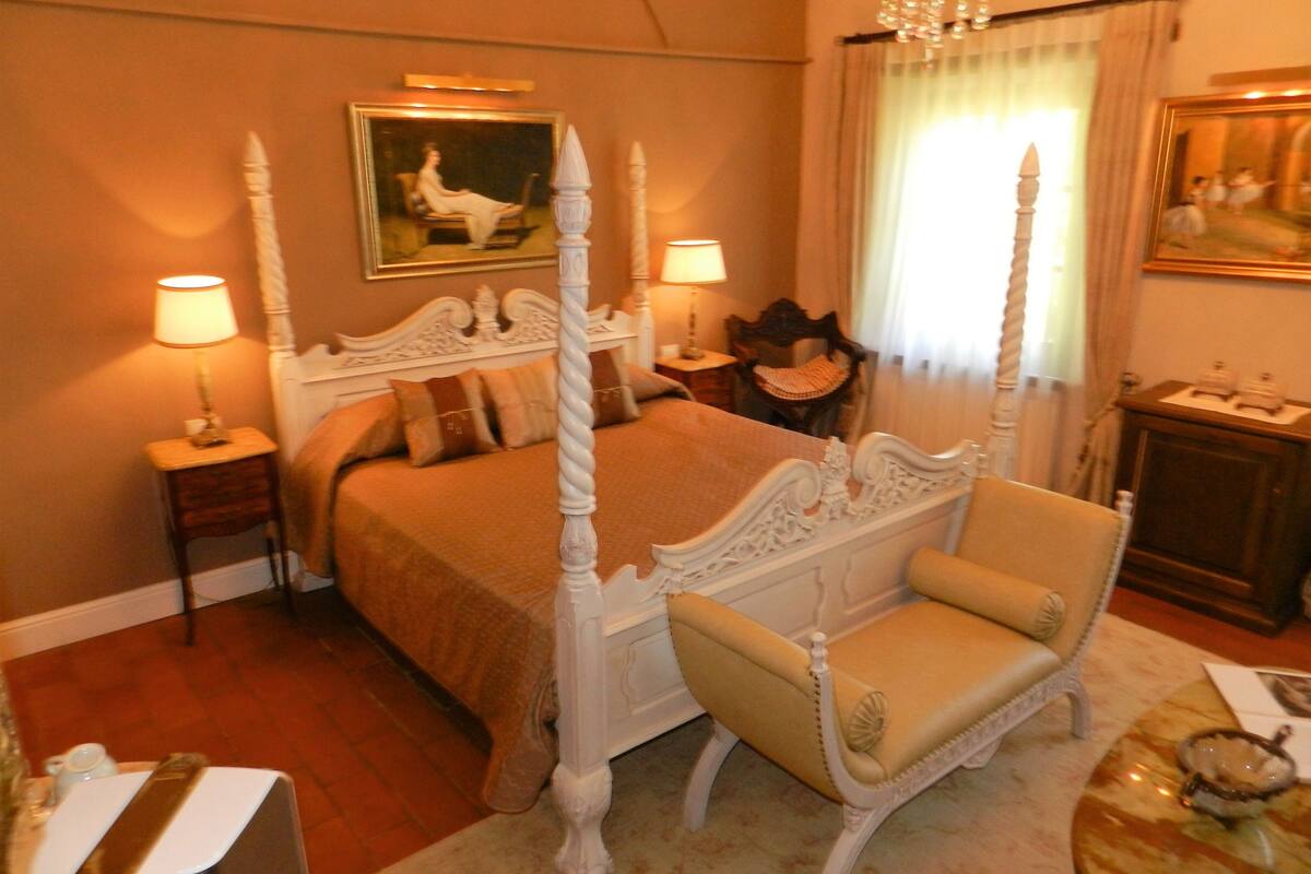 Ein Bett mit Baldachin, ein Kopfteil, zwei Nachttische mit Lampen, ein Sessel, ein Bild an der Wand und ein Fenster mit Vorhängen.