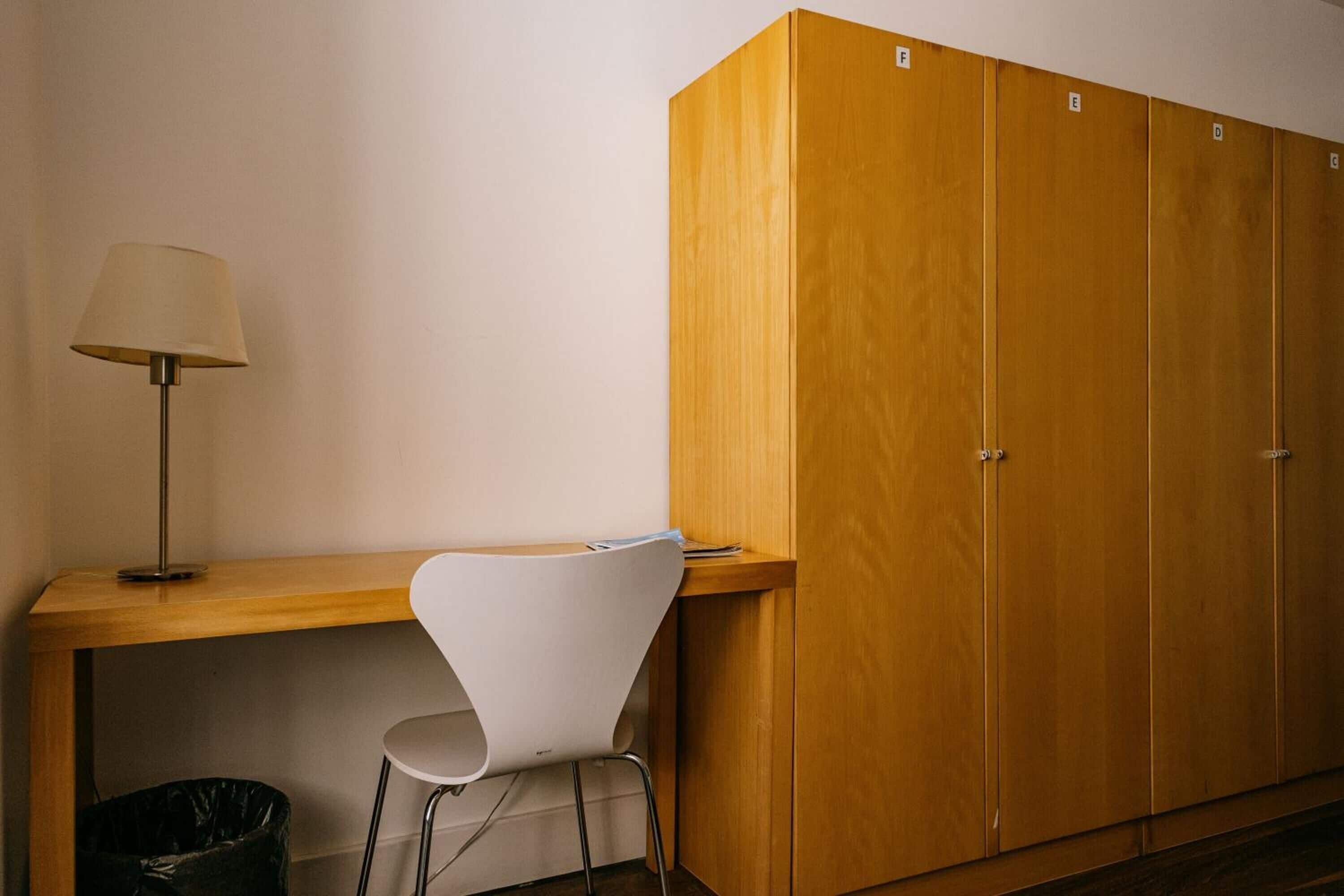 Una habitación con un escritorio de madera, una silla blanca, una lámpara y un armario.