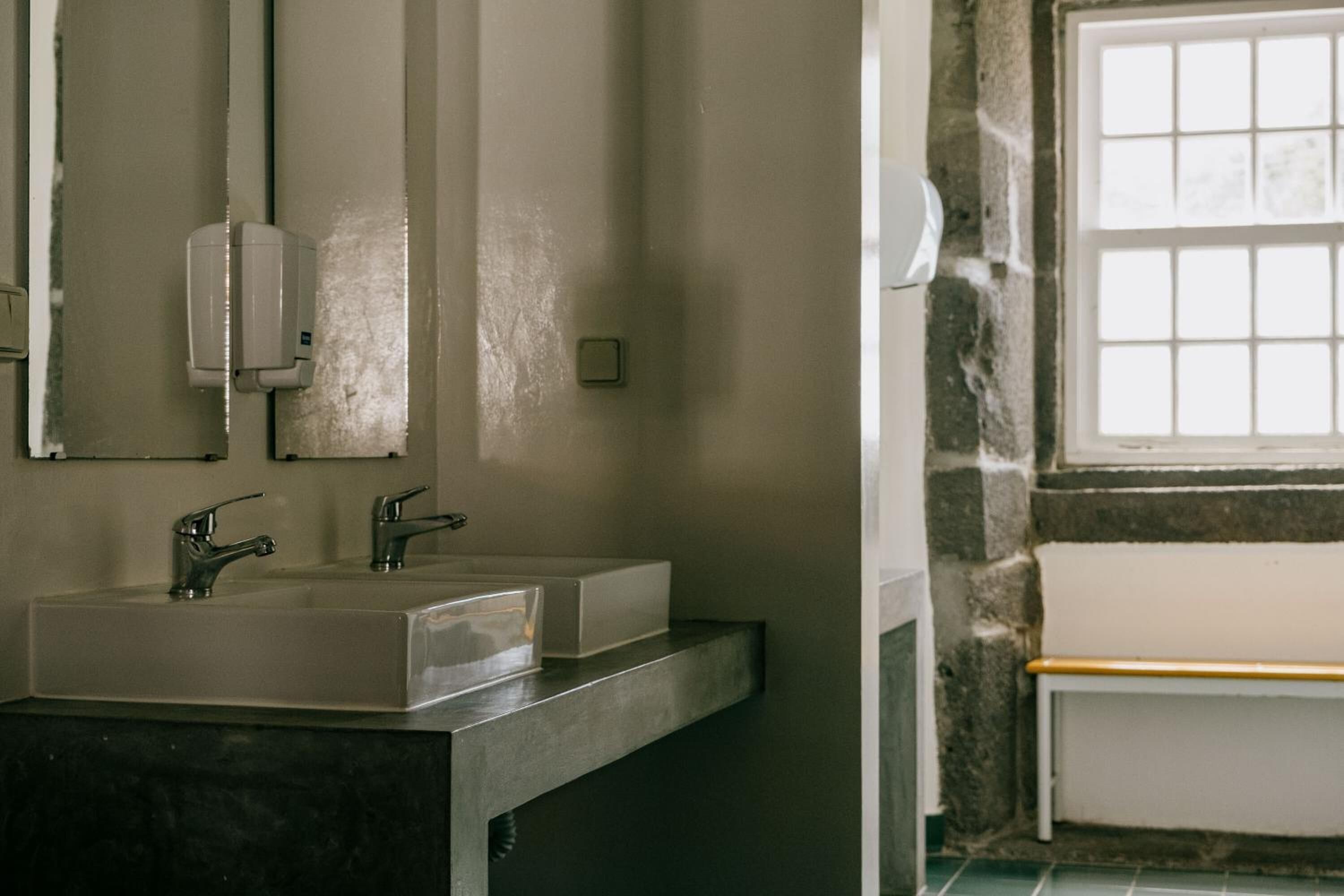 Un baño con lavamanos, dos grifos, dispensador de jabón, espejo y ventana con vistas.