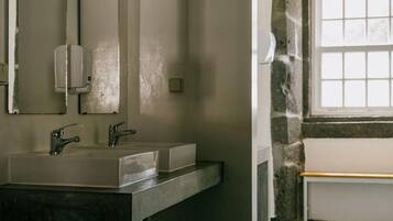 Un baño con lavamanos, dos grifos, dispensador de jabón, espejo y ventana con vistas.