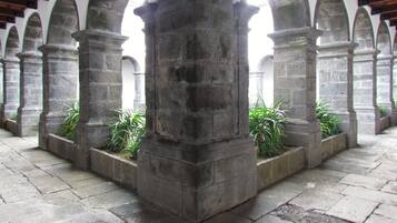 Un corredor de piedra con arcos y columnas, con plantas en macetas y suelo de baldosas.