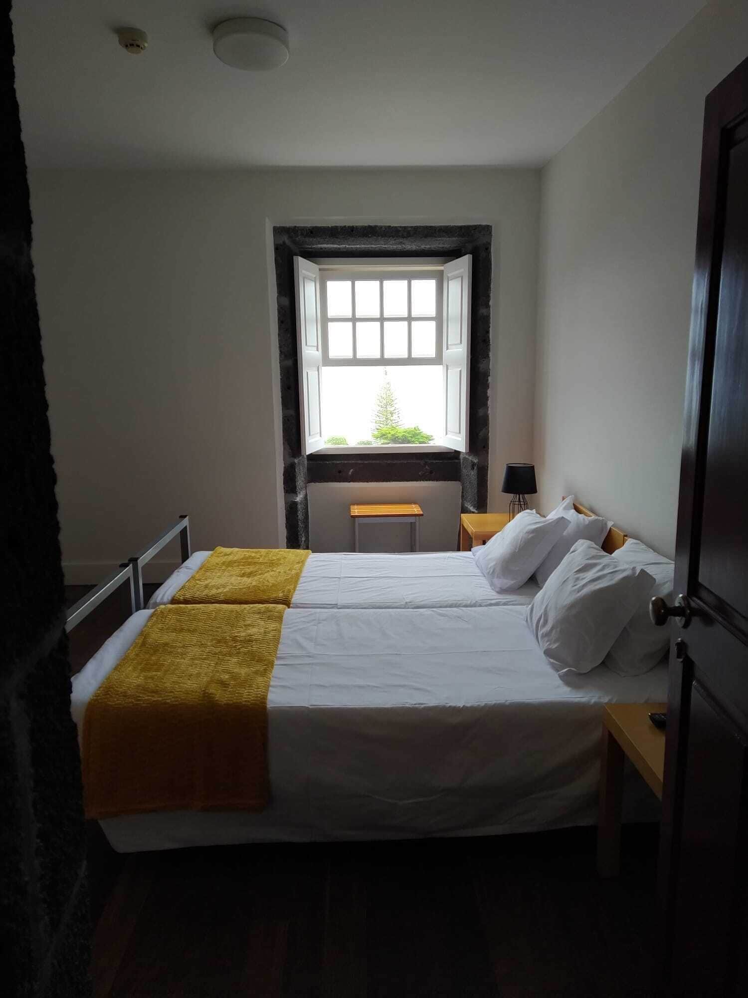 A bedroom with a bed, a nightstand, a lamp, and a window with a view of greenery.