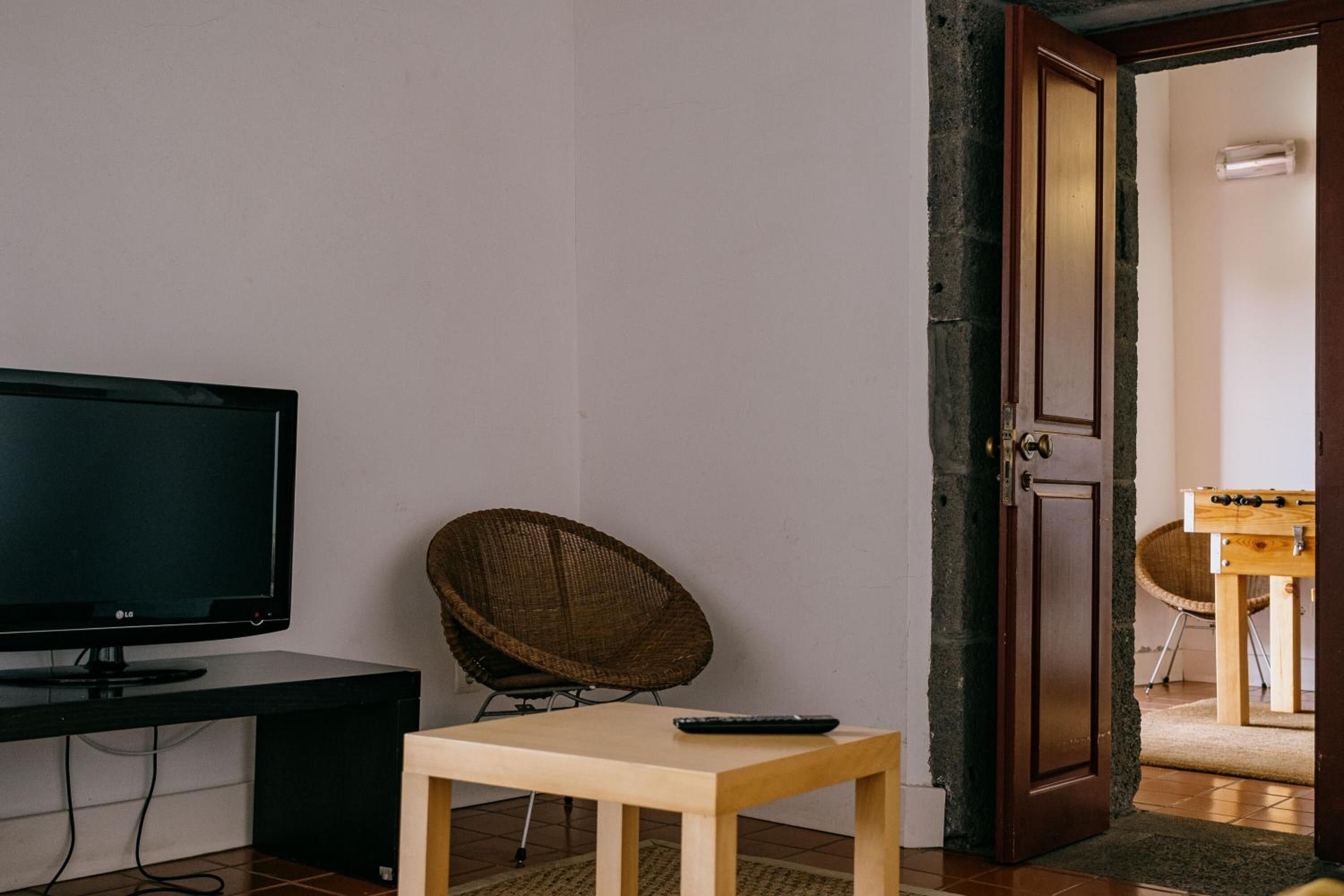 A room with a flat-screen TV on a black stand, a wicker chair, a small wooden table, and a wooden door leading to another room with a foosball table.