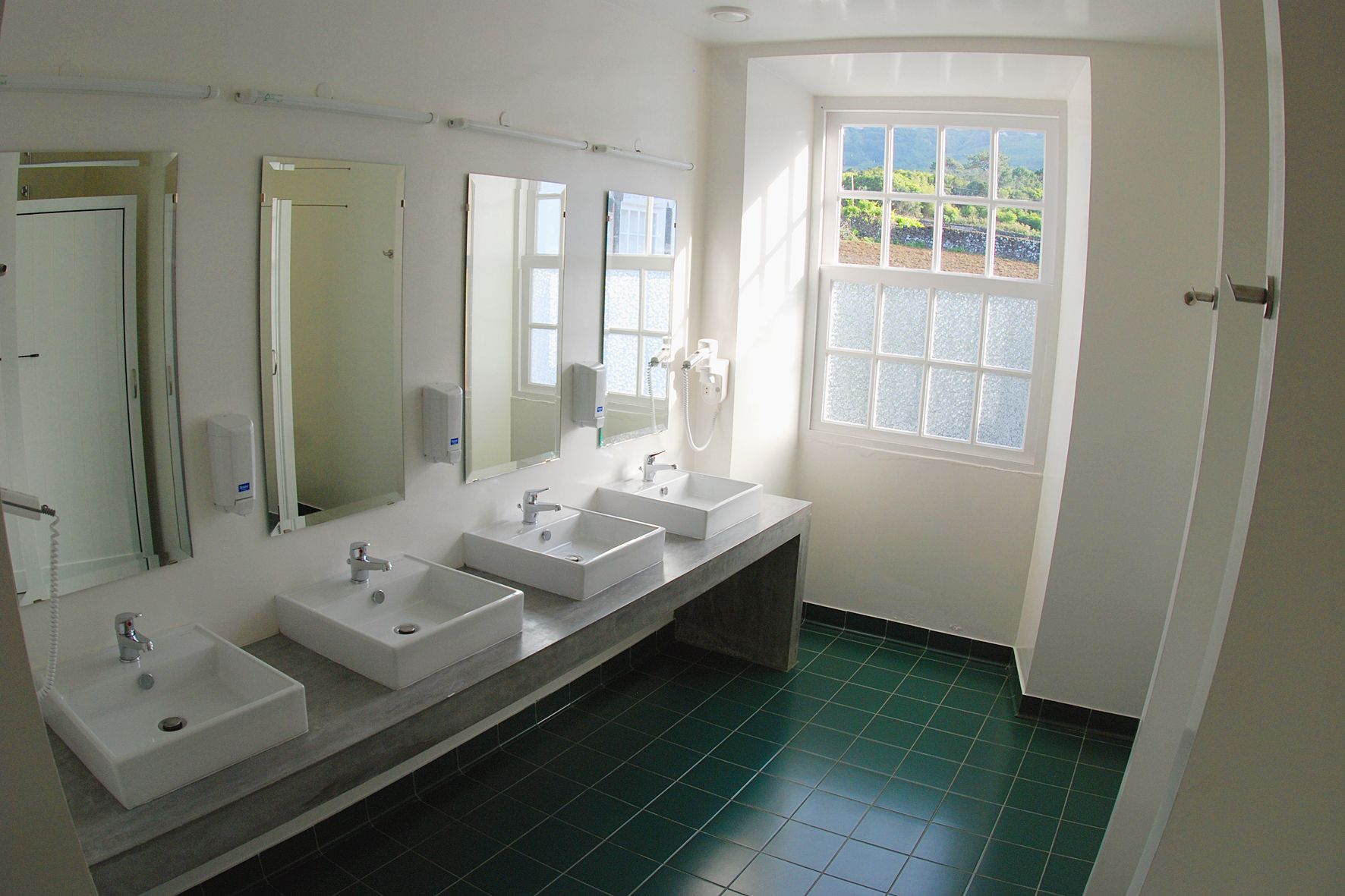 A bathroom with three sinks, a large mirror, and a window with a view of greenery.