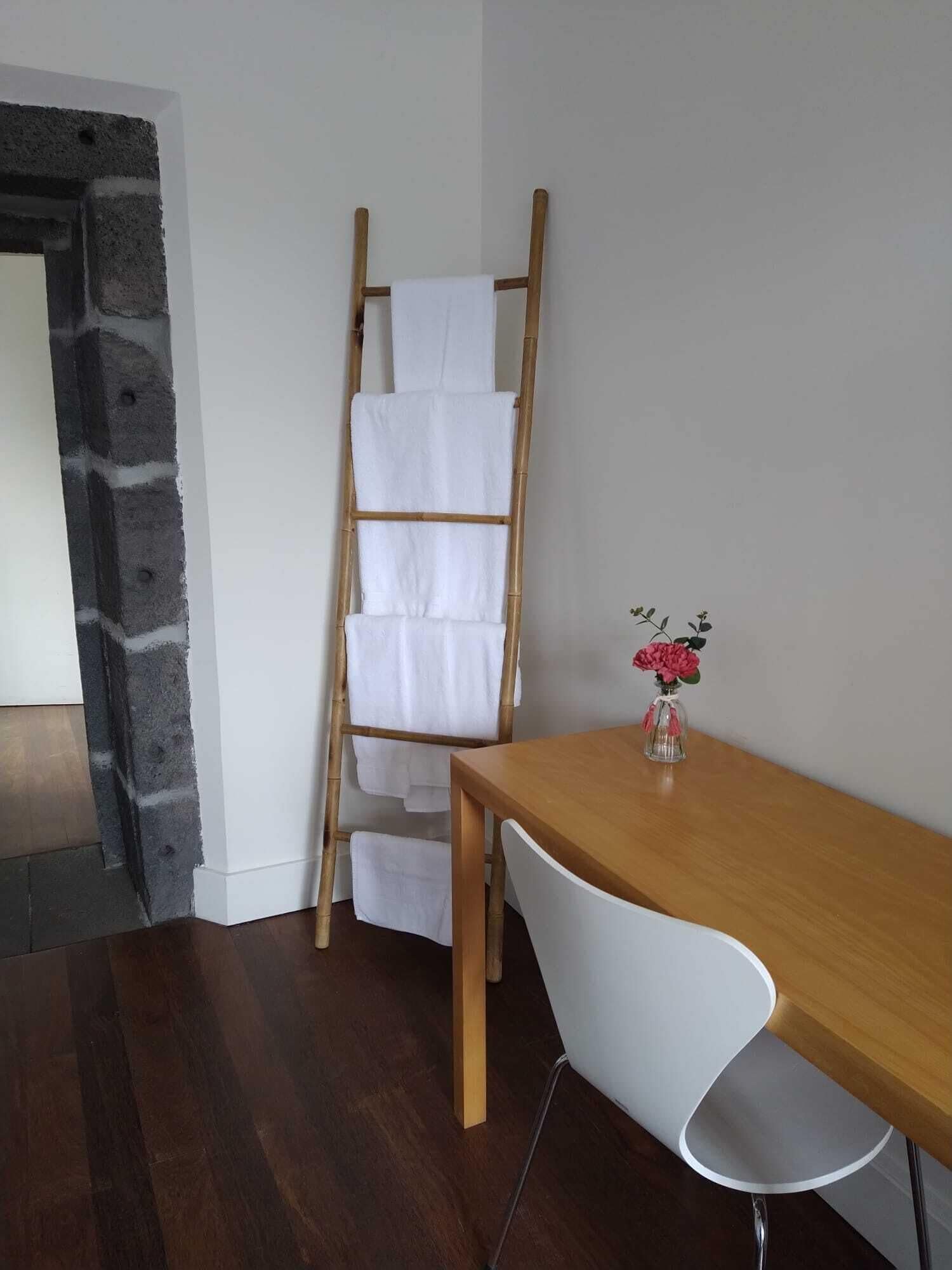 Una habitación con una escalera de madera apoyada en una pared blanca, una mesa de madera, una silla blanca y un pequeño jarrón con flores.