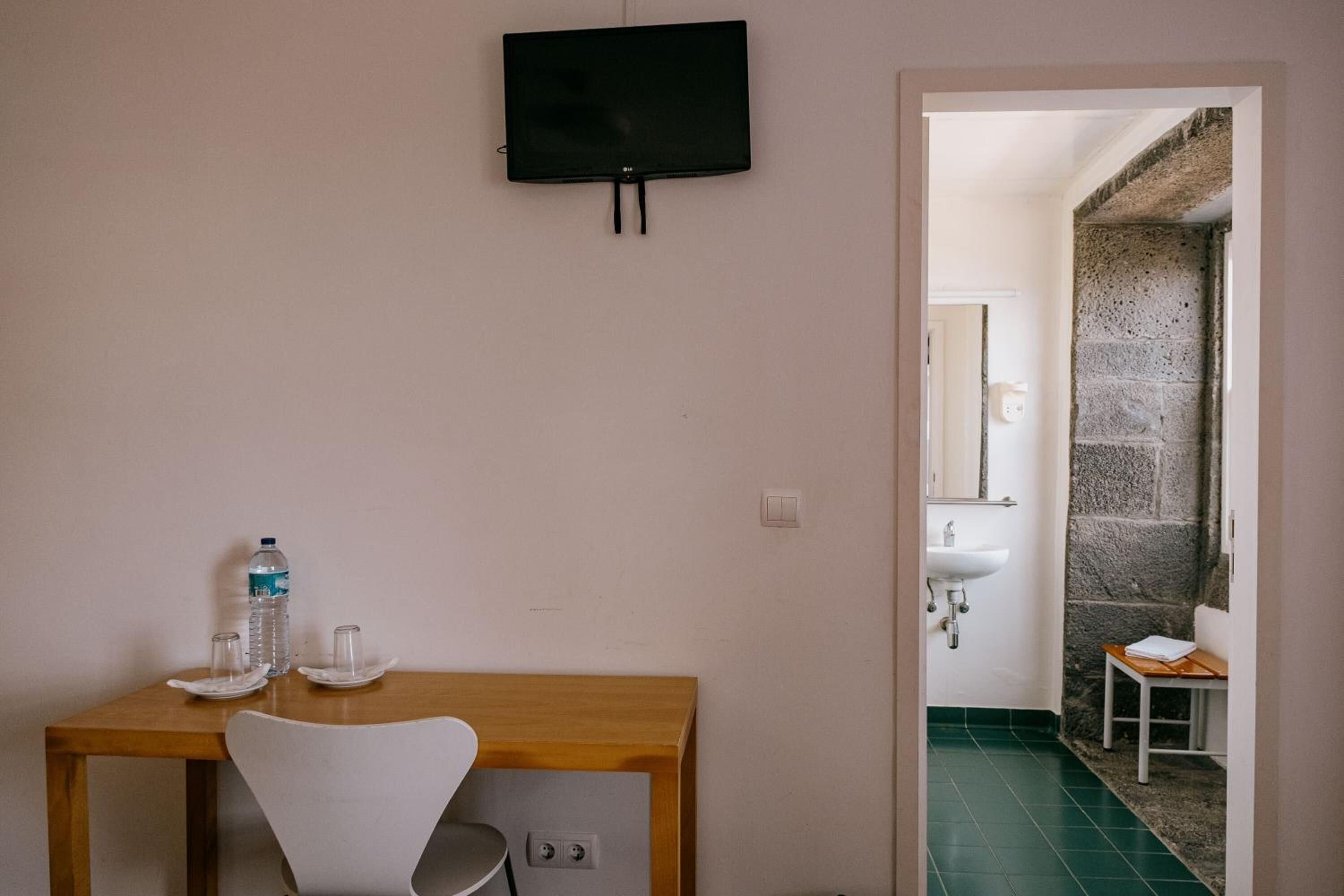 A room with a wooden desk, a chair, a television mounted on the wall, a water bottle, glasses, and a small table with a towel.