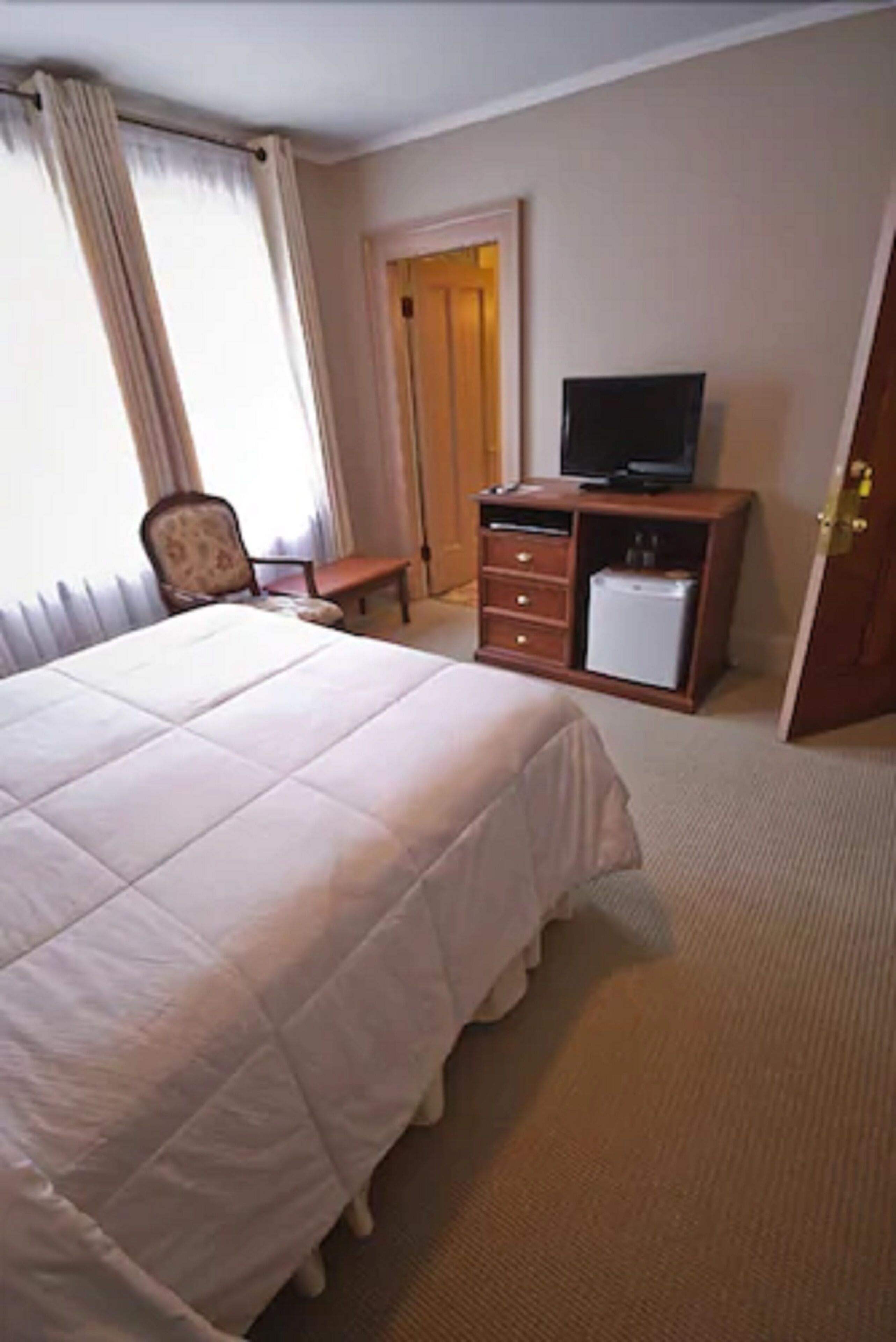 Une chambre d’hôtel avec un grand lit, un téléviseur posé sur une commode en bois, une chaise et une porte donnant sur une autre pièce.
