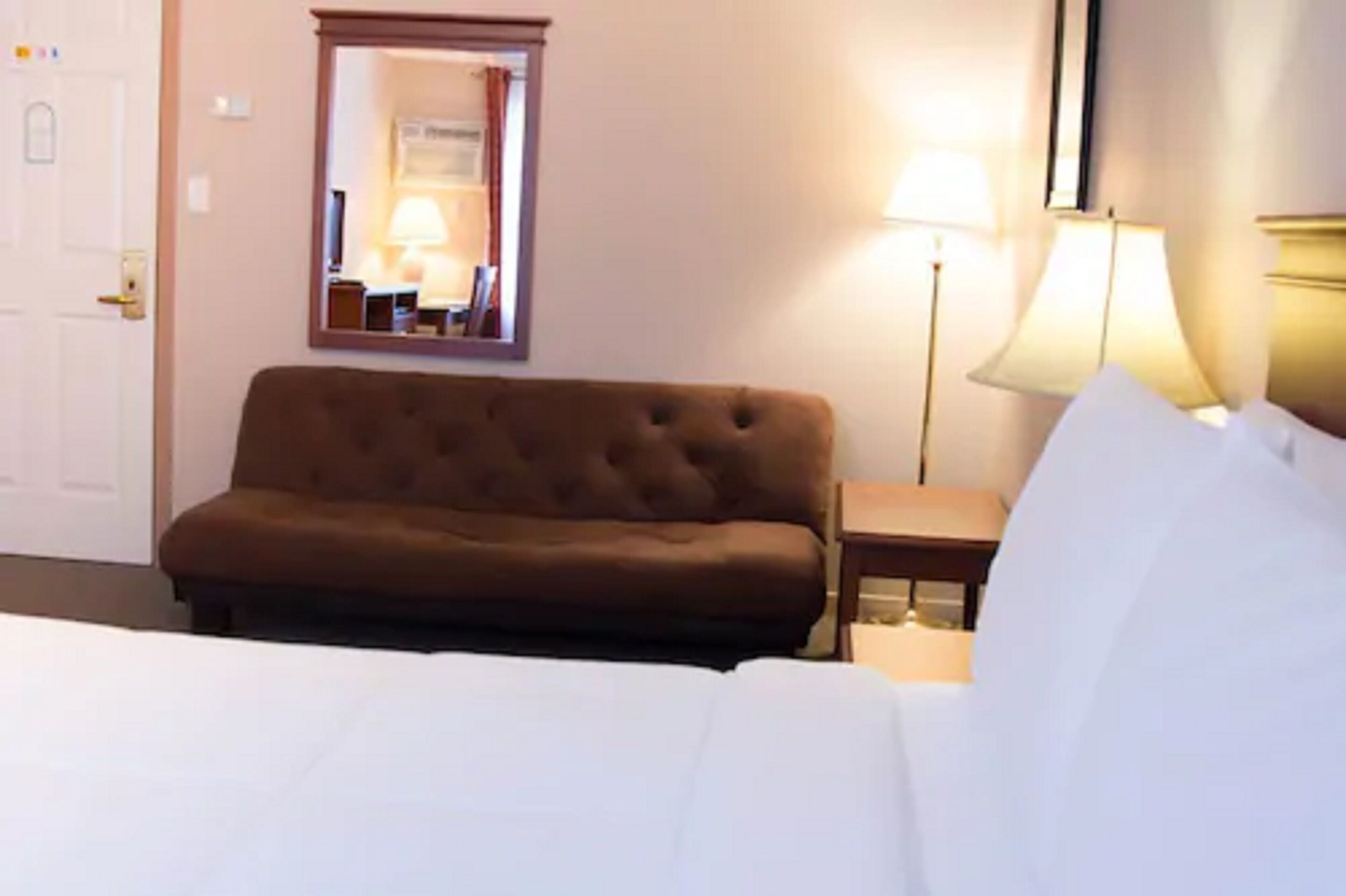 A hotel room with a bed, a brown sofa, a nightstand with a lamp, and a mirror on the wall.