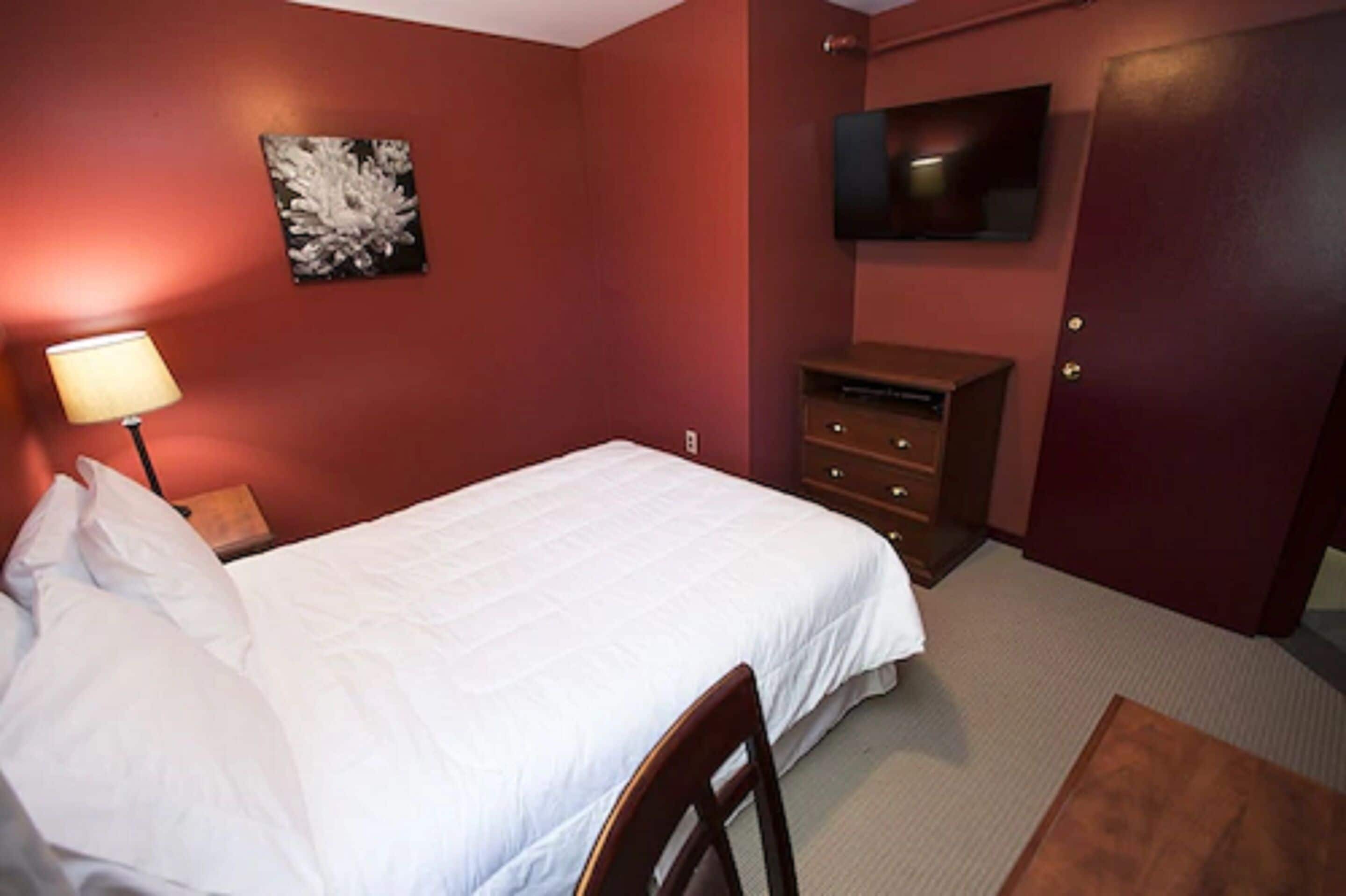 A hotel room with a bed, a desk, a lamp, a dresser, a television, and a door.