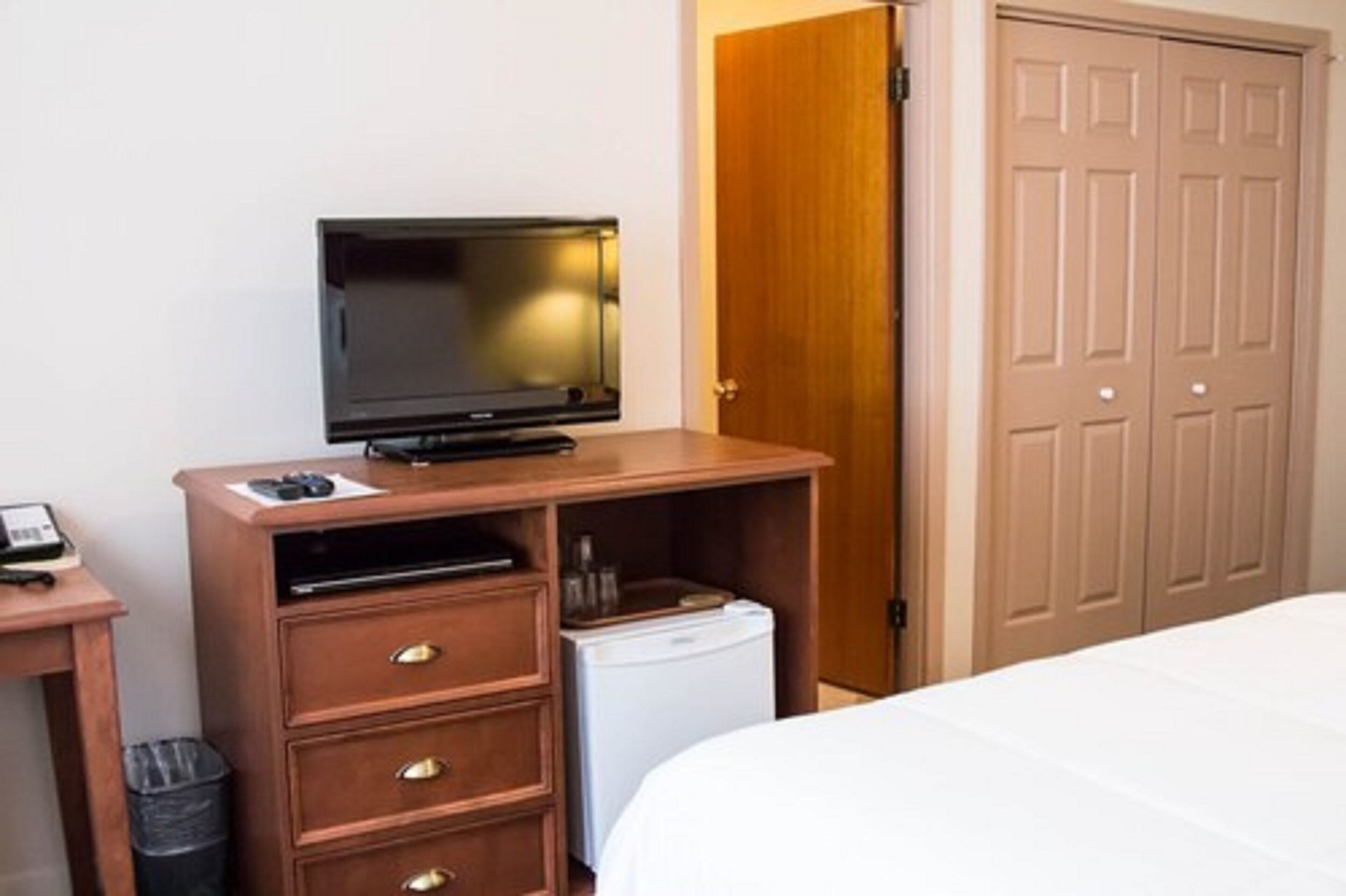 A hotel room with a bed, a wooden desk with a television, a mini-fridge, and a wardrobe.